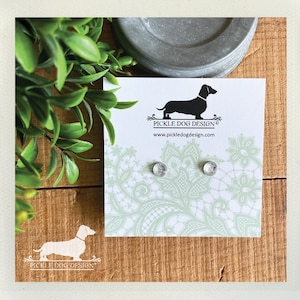 May include: A pair of silver stud earrings on a card with a dachshund logo. The card has a light green floral pattern and the text "PICKLE DOG DESIGN" and "www.pickledogdesign.com". The background is a wooden surface with green leaves.