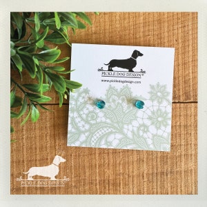 May include: A pair of teal-colored stud earrings on a white card with a dachshund logo and the text "Pickle Dog Design". The card has a light green lace pattern. The earrings are displayed on a wooden surface with greenery.