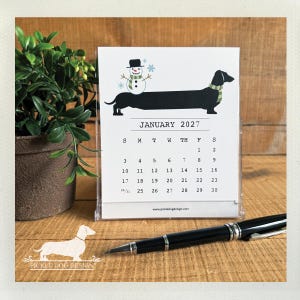 May include: A January 2027 desk calendar in a clear plastic stand featuring a black dachshund wearing a green scarf with a snowman on its back. The calendar page displays the month, year, and dates, with the text www.pickledogdesign.com at the bottom.