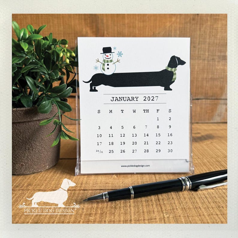 May include: A January 2027 desk calendar in a clear plastic stand featuring a black dachshund wearing a green scarf with a snowman on its back. The calendar page displays the month, year, and dates, with the text www.pickledogdesign.com at the bottom.