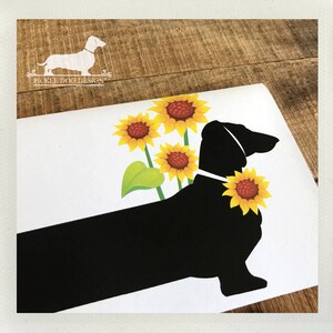 May include: A white card featuring a black dachshund silhouette with yellow sunflowers. The dog has a sunflower collar and is positioned in front of three sunflowers. The top left corner has the text "Pickle Dog Design" with a dachshund logo.