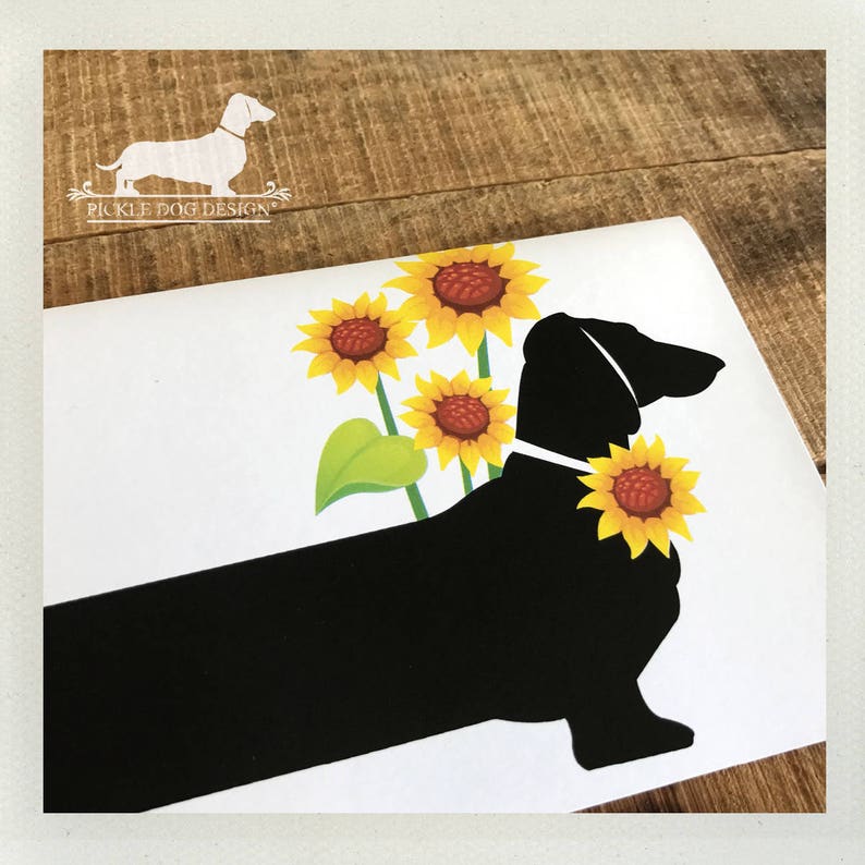 May include: A white card featuring a black dachshund silhouette with yellow sunflowers. The dog has a sunflower collar and is positioned in front of three sunflowers. The top left corner has the text "Pickle Dog Design" with a dachshund logo.