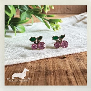 May include: A pair of cherry-shaped stud earrings. Each earring features two pink, gem-like cherries with green enamel leaves. The earrings are set against a neutral fabric and wooden background, with a sprig of green leaves in the upper left corner.