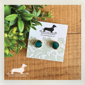 May include: A pair of round, teal-colored stud earrings with gold-colored rims. The earrings are displayed on a white card with a dachshund logo and the text "Pickle Dog Design". The background is a wooden surface with green foliage.