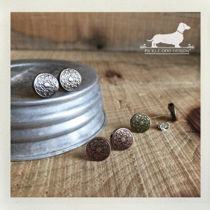 May include: A collection of round stud earrings in various metallic finishes, including silver, bronze, and copper. Each earring features a detailed, embossed floral design. The earrings are displayed on a wooden surface.