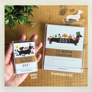 May include: A mini and a standard size 2027 12-month calendar in clear plastic cases. The calendars feature a black dachshund illustration with seasonal icons, the text 2027, 12-MONTH CALENDAR, and pickledogdesign.com. A brown paper band wraps around each.