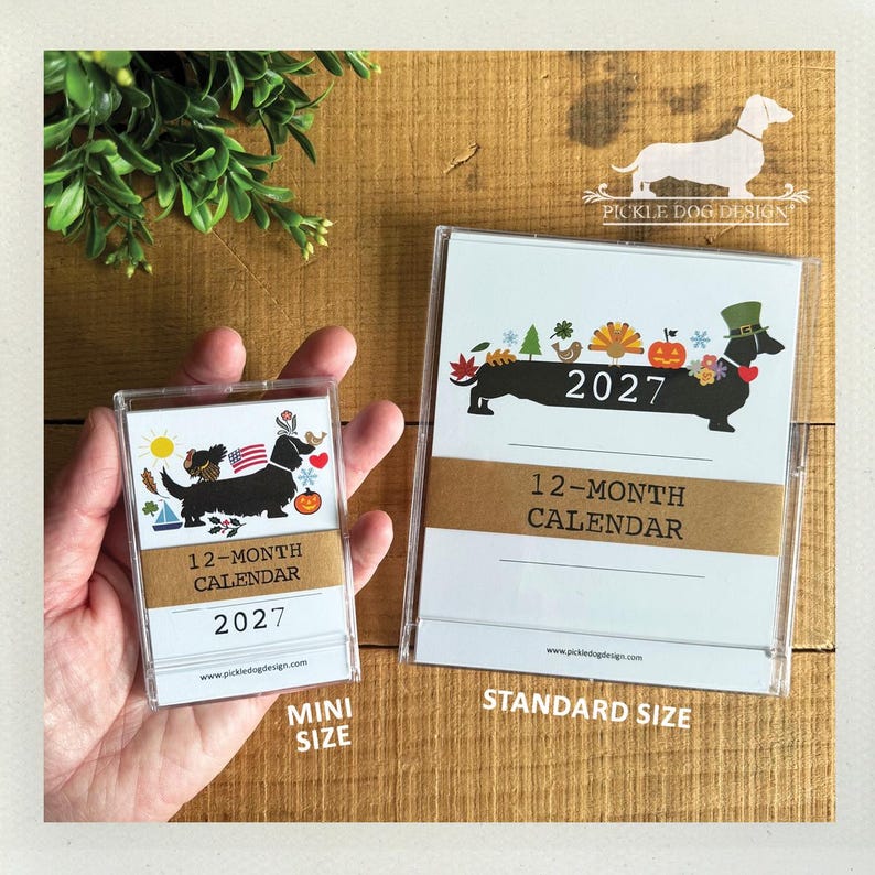 May include: A mini and a standard size 2027 12-month calendar in clear plastic cases. The calendars feature a black dachshund illustration with seasonal icons, the text 2027, 12-MONTH CALENDAR, and pickledogdesign.com. A brown paper band wraps around each.