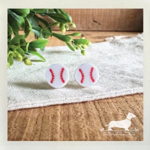 May include: A pair of white baseball stud earrings with red stitching details. The earrings are round and sit on a white cloth, with greenery and a wooden background. The Pickle Dog Design logo is in the bottom right corner.