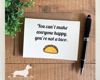 May include: A greeting card with the text "You can't make everyone happy, you're not a taco." and a taco illustration. The card is on a wooden surface with a pen and greenery. The Pickle Dog Design logo is visible.