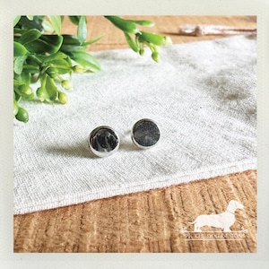 May include: Two silver-colored, round cufflinks with a reflective surface are displayed on a white linen cloth. The cufflinks are set against a backdrop of greenery and a wooden surface, suggesting a handcrafted or artisanal product.