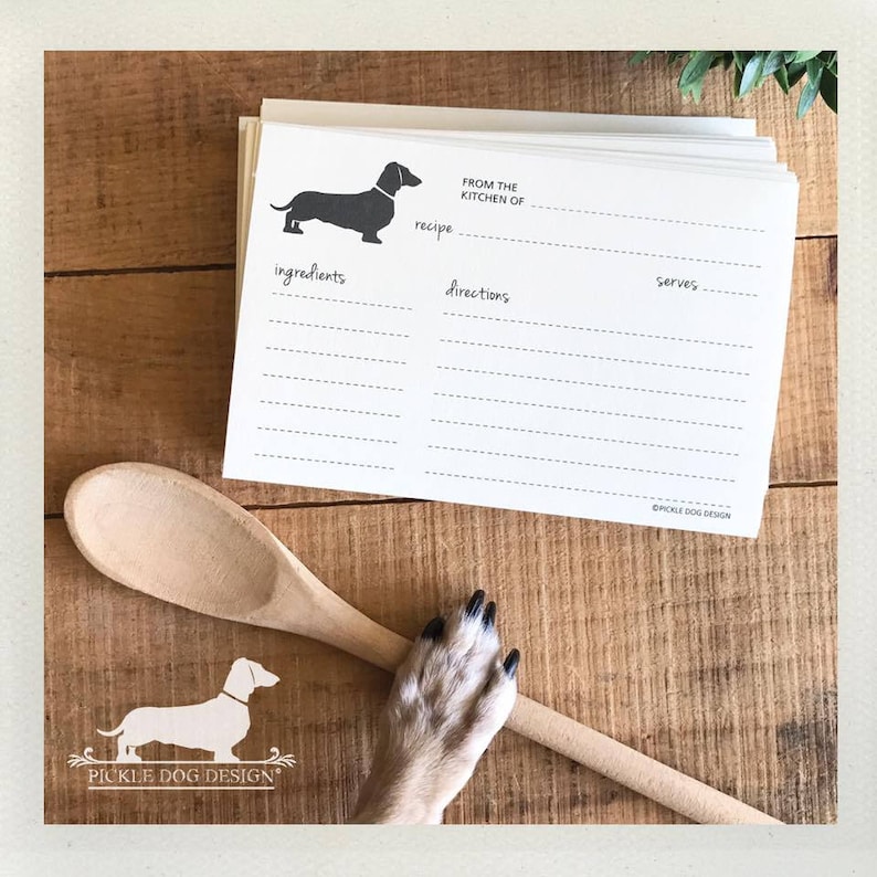 May include: A stack of white recipe cards with a dachshund silhouette and the text "From the Kitchen of." A wooden spoon and a dog's paw rest on a wooden surface. The logo "Pickle Dog Design" is visible.