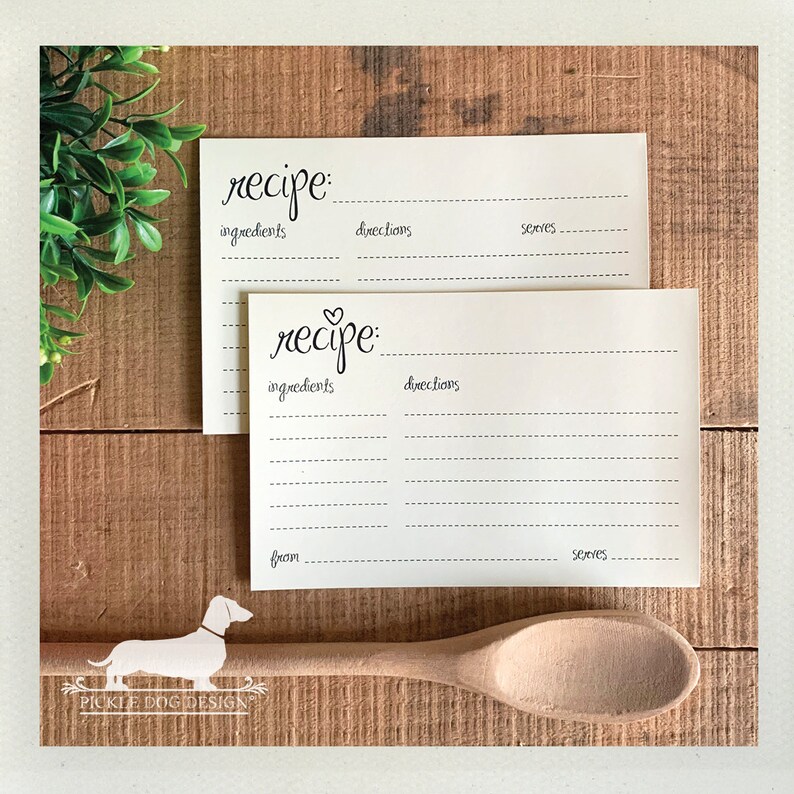 May include: Two cream-colored recipe cards with handwritten "recipe" text and dotted lines for ingredients, directions, and servings. A wooden spoon and a green plant are also in the image. The cards are on a wooden surface.