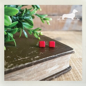 May include: A pair of small, square, bright red stud earrings. The earrings are displayed on a vintage book with a distressed cover, alongside green foliage. The background features a wooden surface and the Pickle Dog Design logo.