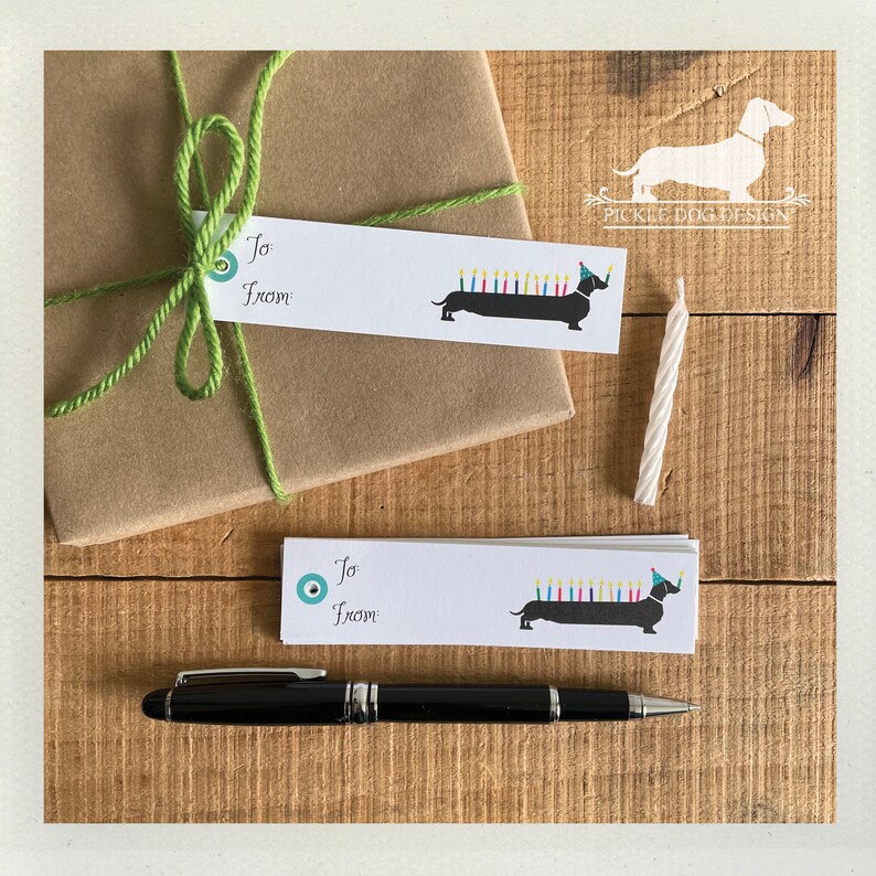 Pu&ograve; includere: Un regalo avvolto in carta marrone con un fiocco di spago verde. Due etichette regalo bianche con un bassotto con cappello da festa e candele. Una candela di compleanno bianca e una penna nera su legno.