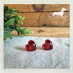 以下が含まれることがあります： キラキラした質感の赤いハート型のクリスタルイヤリング2つ。オフホワイトの質感のある表面に展示されています。背景には木の板の背景と緑の葉の小枝があります。