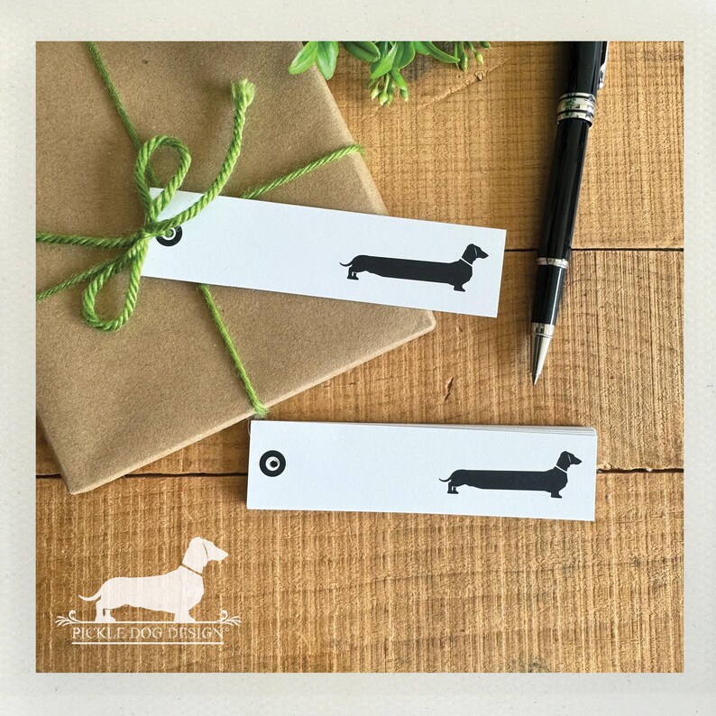 Pu&ograve; includere: Un regalo avvolto in carta marrone legato con spago verde, accompagnato da due etichette rettangolari bianche. Ogni etichetta presenta una silhouette nera di bassotto e un cerchio nero. Una penna nera poggia su una superficie di legno.