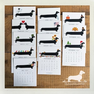 May include: A 2027 wall calendar set featuring twelve individual white cards, each displaying a black silhouette of a dachshund with seasonal illustrations for every month, including holiday themes, flowers, and a calendar grid.