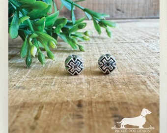 May include: A pair of silver-colored stud earrings with a geometric cross design. The earrings are round and have a detailed, openwork pattern. The background features green foliage and a wooden surface.