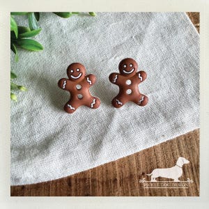 May include: Two brown gingerbread man-shaped buttons with white icing details, including a smiling face, buttons, and decorative edges. The buttons are set on a light-colored fabric, with a wooden surface below. The image includes a logo for Pickle Dog Design.