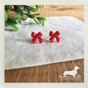 May include: A pair of bright red bow-shaped earrings. The earrings are positioned on a light gray linen surface, with green foliage in the background. The earrings are a glossy red color and are a cute accessory.