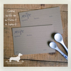 May include: Two recipe cards with the word "recipe" in cursive, with lines for ingredients, directions, and servings. The cards are on a wooden surface with two white and blue measuring spoons. Text reads "choose with or without a heart."