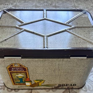 May include: A rectangular metal bread box with a dark brown lid featuring a geometric diamond pattern. The beige body has a textured finish and displays a graphic illustration of a scale with the text Weights & Measures and the word Bread.