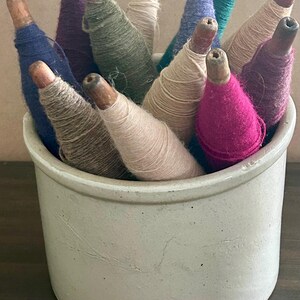 May include: A collection of wooden bobbins wrapped in various colors of yarn, including blue, green, beige, purple, and magenta, standing upright inside a plain, off-white ceramic crock with a smooth, cylindrical shape and a slightly textured surface.