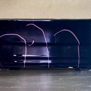 May include: A rectangular ceramic planter with a glossy black finish and a series of thin, vertical pink lines that curve across the surface. The planter features a stepped, tiered design along the sides and a wide, flared rim at the top.