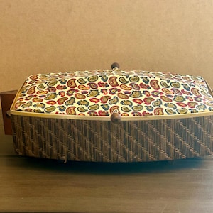 May include: A rectangular sewing basket with a paisley patterned lid in red, yellow, and blue. The basket has a woven brown exterior and wooden accents. The lid is secured with a wooden knob closure.