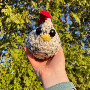 May include: A handmade plush chicken with a red comb, black eyes, and a yellow beak. The chicken is primarily white with black speckles. The background is a blurred view of green foliage and blue sky.