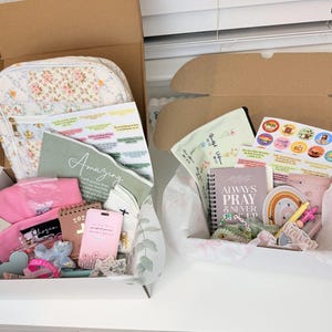Peut inclure: Deux coffrets cadeaux remplis de divers articles. L'un contient un sac &agrave; motifs floraux, des v&ecirc;tements roses et de la papeterie. L'autre comprend un carnet avec le texte "Always Pray & Never Give Up", des autocollants et une d&eacute;coration arc-en-ciel. Les deux bo&icirc;tes sont dans des bo&icirc;tes en carton marron.