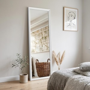 Peut inclure: Un grand miroir à cadre blanc est appuyé contre un mur de couleur neutre. Le miroir reflète un panier en osier, une plante en pot et une fenêtre. Une impression encadrée et un lit avec une literie grise sont également visibles.