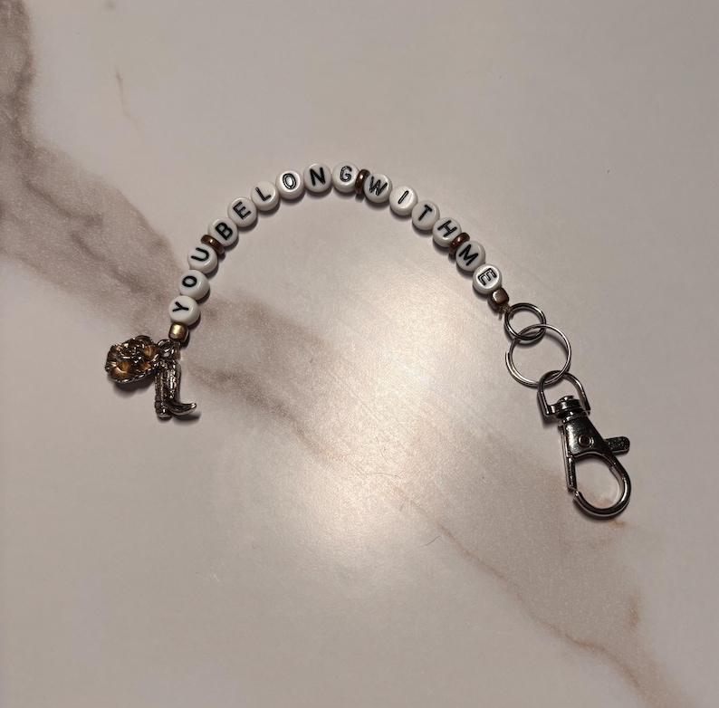 May include: A beaded keychain with the phrase "YOU BELONG WITH ME" spelled out in black letters on white beads. It features a small gold-coloured charm and a silver-coloured clasp.