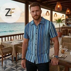 May include: Short-sleeved button-down shirt with a blue and white geometric pattern. The shirt has a camp collar and is worn with black pants. The image is taken outdoors, with a restaurant and ocean in the background.