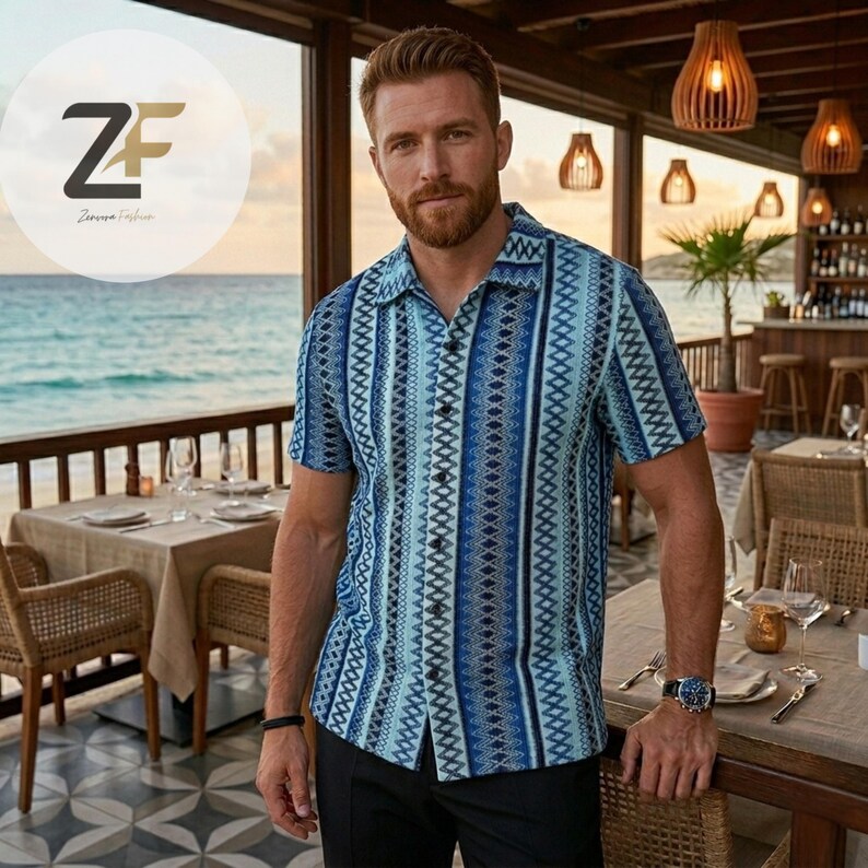 May include: Short-sleeved button-down shirt with a blue and white geometric pattern. The shirt has a camp collar and is worn with black pants. The image is taken outdoors, with a restaurant and ocean in the background.