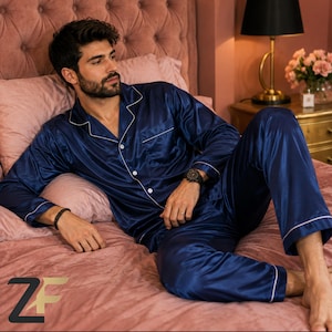 May include: A navy blue satin pajama set featuring a long-sleeved button-down shirt with a notched collar and matching trousers. The pajamas are detailed with white piping along the collar, front placket, and sleeve cuffs, creating a classic look.