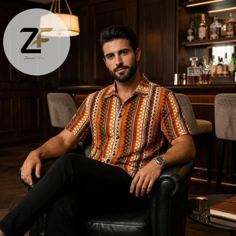 May include: Short-sleeved button-down shirt with a vertical geometric pattern in shades of brown, orange, and white. The shirt is worn with black pants. The image features a man sitting in a black leather chair.
