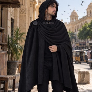 May include: A black hooded cloak featuring a textured fabric, a deep hood, and a circular fastener at the shoulder. The garment drapes over the shoulders and chest, extending downwards in long, flowing folds, providing a full-length silhouette.