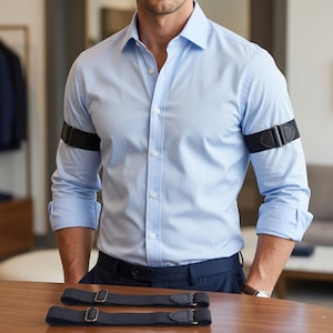May include: A light blue button-down shirt with rolled sleeves held in place by black elastic sleeve garters. The garters feature metal adjusters and leather-like tabs. Two additional black elastic sleeve garters are placed on the wooden surface.