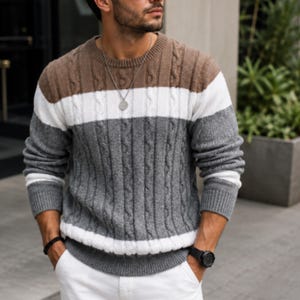 May include: A long-sleeve crew neck sweater featuring a color-blocked design with horizontal stripes in brown, white, and gray. The knit fabric displays a vertical cable knit pattern throughout the body and sleeves, finished with ribbed cuffs.