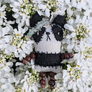 May include: A handmade panda bear doll with black and white stitched details, wooden bead arms and legs, and a green beaded headband. The doll is surrounded by a bed of white flowers.