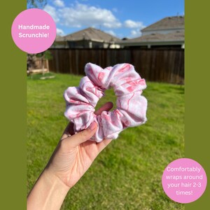 May include: A pink and white patterned fabric scrunchie with a gathered, ruffled texture. The scrunchie features the text Handmade Scrunchie! and Comfortably wraps around your hair 2-3 times! printed on pink circular overlays on the image.