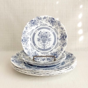 May include: A stack of white ceramic plates with a blue floral design. The plates have a scalloped edge and a central floral motif. A smaller plate rests on a decorative metal stand, with more plates stacked below.