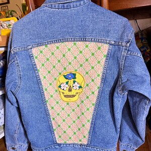 Könnte beinhalten: Hellblaue Jeansjacke mit einem farbenfrohen Totenkopf-Patch auf dem Rücken. Der Totenkopf ist gelb mit Blumenakzenten und ist auf einem gemusterten Stoffpaneel zentriert. Das Panel hat ein rosa-grünes Design.