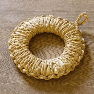 May include: A circular trivet, woven from natural straw, with a central hole and a small loop for hanging. The straw is tightly woven, creating a textured surface. This kitchen accessory is designed to protect surfaces from hot items. The diameter is approximately 20 cm.