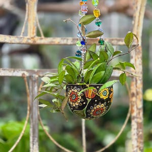Puede incluir: Una maceta colgante con base negra y un patrón floral colorido en rojo, amarillo y verde. La maceta contiene una planta de hojas verdes y está suspendida por una cadena plateada adornada con cuentas de vidrio multicolor de varias formas.