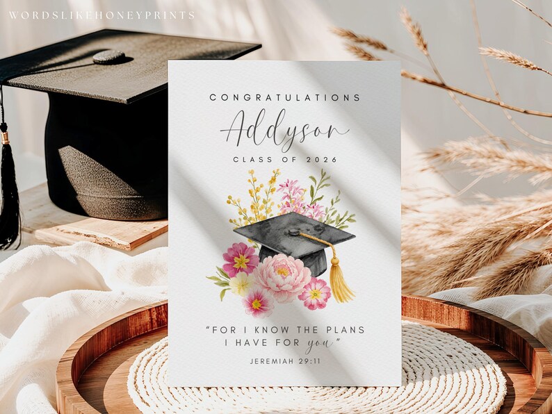 Puede incluir: Tarjeta de graduaci&oacute;n con el nombre "Addyson" y "Clase de 2026" en escritura elegante. La tarjeta presenta una ilustraci&oacute;n de acuarela de un birrete, flores rosas y la cita de Jerem&iacute;as 29:11.