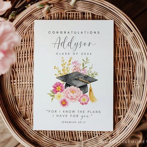 Puede incluir: Tarjeta de graduaci&oacute;n con ilustraci&oacute;n floral y birrete. La tarjeta dice "Felicitaciones Addyson Clase de 2026" e incluye la cita "Porque yo s&eacute; los planes que tengo para vosotros" de Jerem&iacute;as 29:11. La tarjeta est&aacute; sobre una cesta tejida.