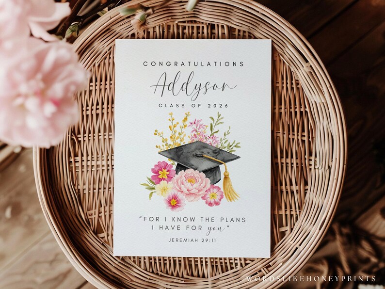 Puede incluir: Tarjeta de graduaci&oacute;n con ilustraci&oacute;n floral y birrete. La tarjeta dice "Felicitaciones Addyson Clase de 2026" e incluye la cita "Porque yo s&eacute; los planes que tengo para vosotros" de Jerem&iacute;as 29:11. La tarjeta est&aacute; sobre una cesta tejida.