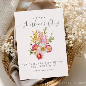 May include: A Mother's Day card with a floral bouquet design. The card features the text "Happy Mother's Day" and a quote from Proverbs 31:28. The bouquet includes pink, yellow, and white flowers with green foliage. The card is set on a woven basket.